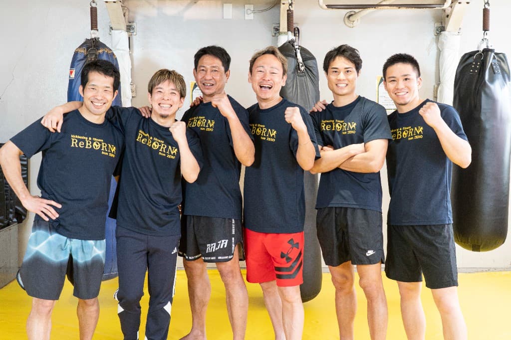 黄色マットのジムフロアに並び、ReBORNロゴのネイビーTシャツで笑顔を見せる男性スタッフ6名の集合写真