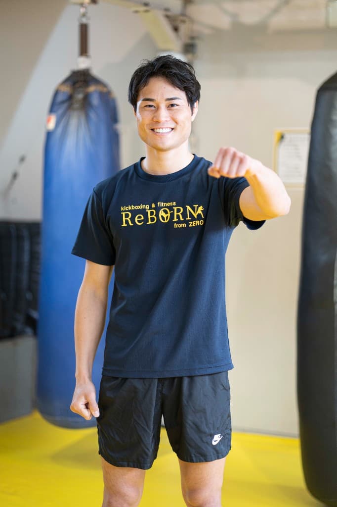 黄色マットのジムで、両脇に青と黒のサンドバッグがある空間で、ReBORNロゴ入りの黒いTシャツ姿に右手のパンチを突き出し笑う池田幸司（現役チャンピオン）