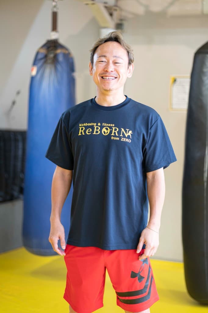 黄色マットのジム内で、両脇のサンドバッグのあいだに立ち、ReBORNロゴ入りのネイビーTシャツで笑顔を見せる代表・斉藤淳