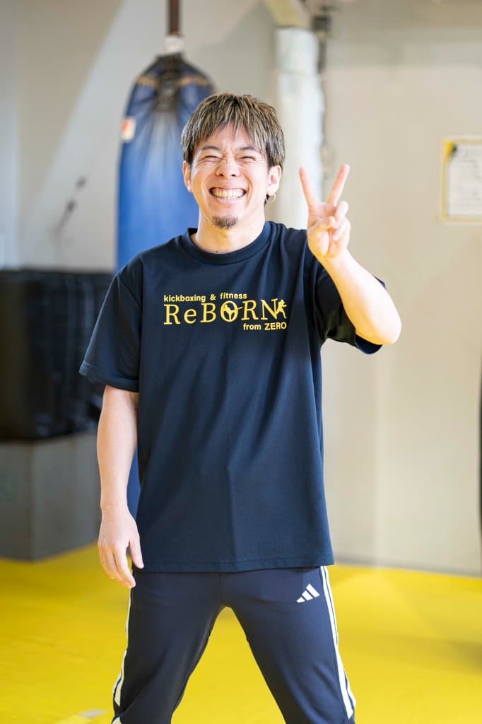 黄色マットのジム内で、ReBORNロゴ入りのネイビーTシャツとトラックスーツ姿で左手をピースにして笑顔を見せる正野宗一郎（プロキックボクサー）